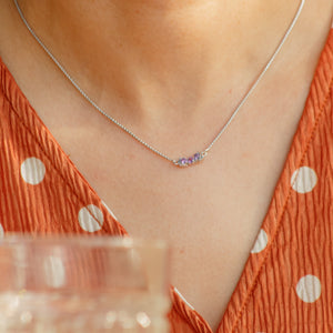 Person wearing a silver necklace with small colored stones, holding a glass.